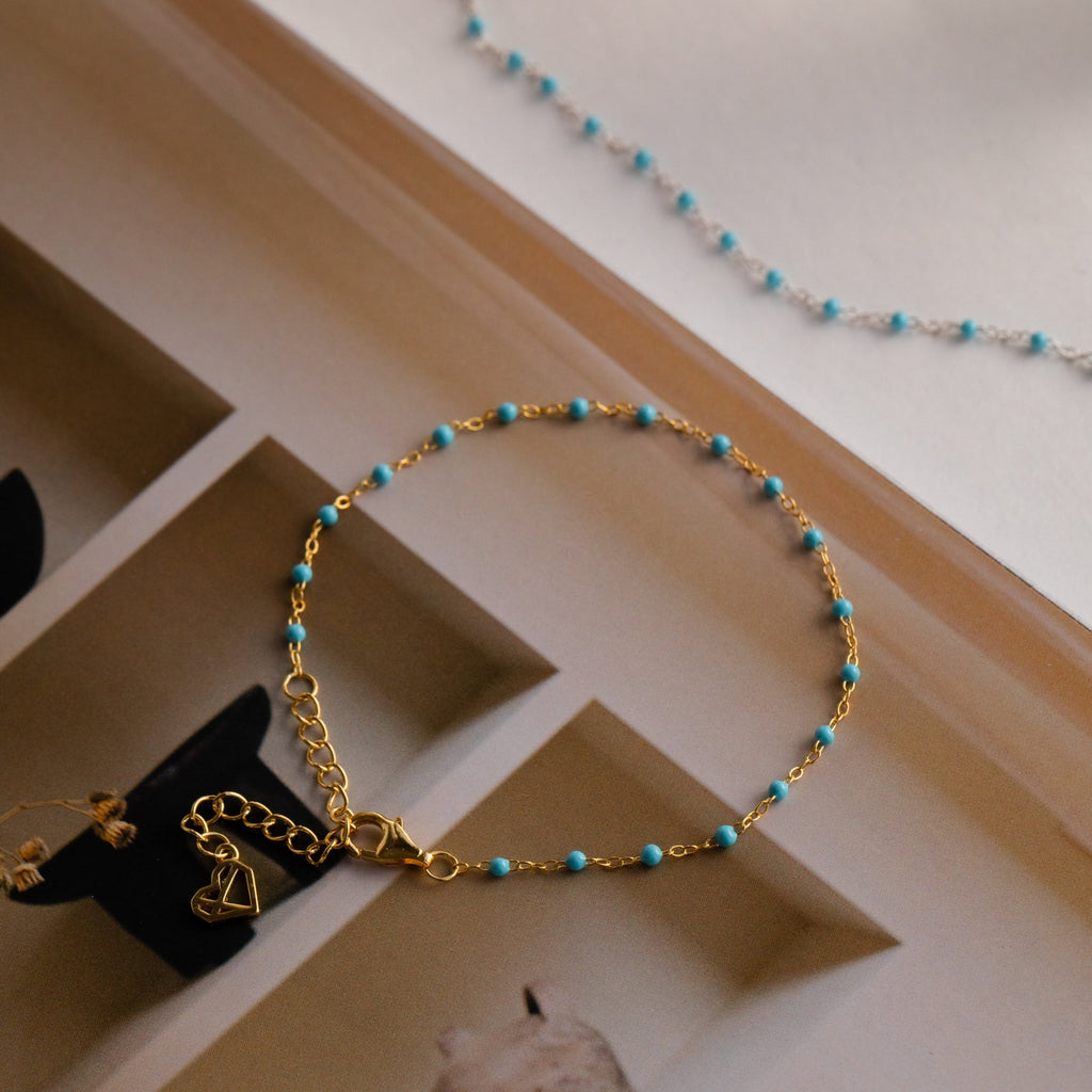The Turquoise Station Bracelet features a gold chain with small turquoise beads, adding a boho touch to your bracelet stack. Shown styled on an open book.