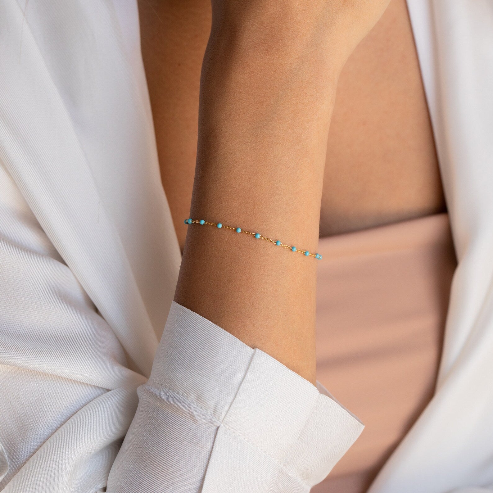 The Turquoise Station Bracelet adds a delicate touch of elegance to her wrist, beautifully complementing her bracelet stack with a white blouse and peach top.