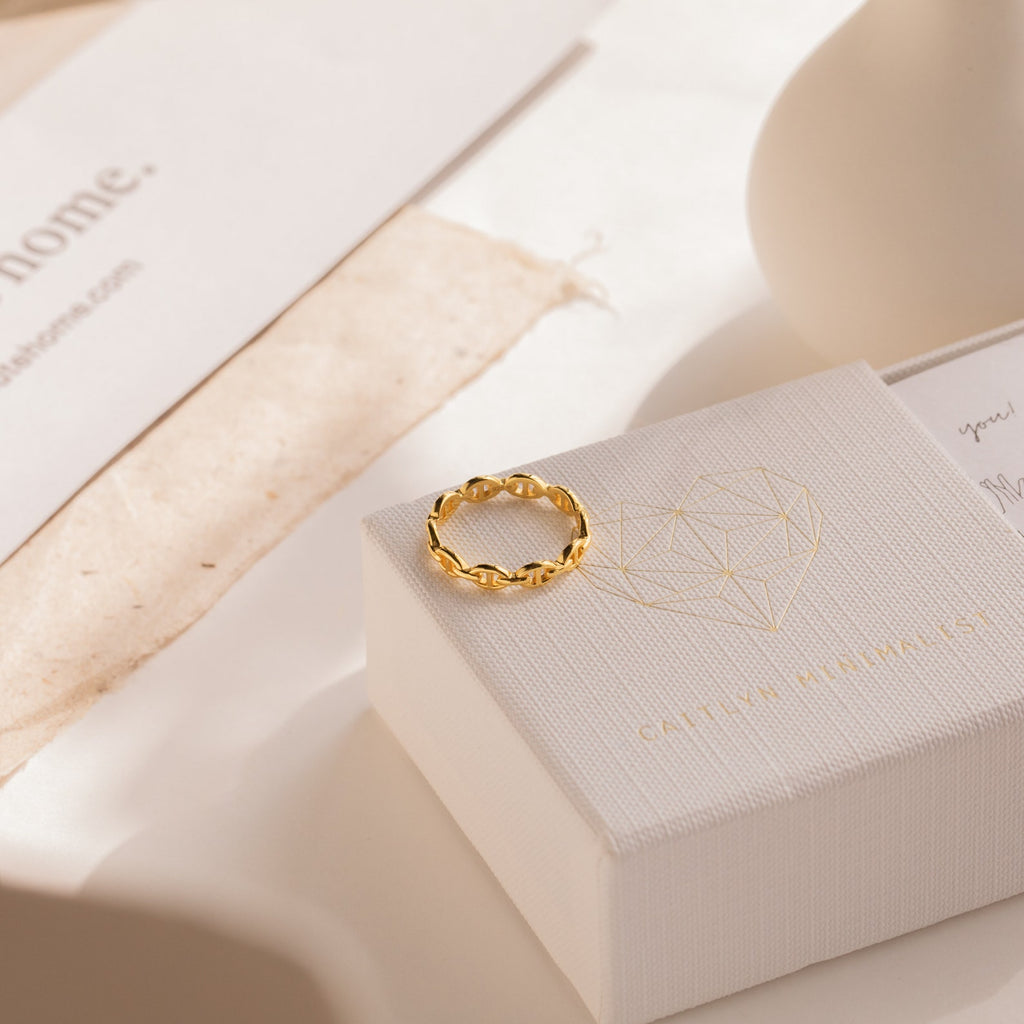 The Reese Link Ring, a gold braided chain design, sits atop a white jewelry box featuring a geometric heart pattern in gentle sunlight.
