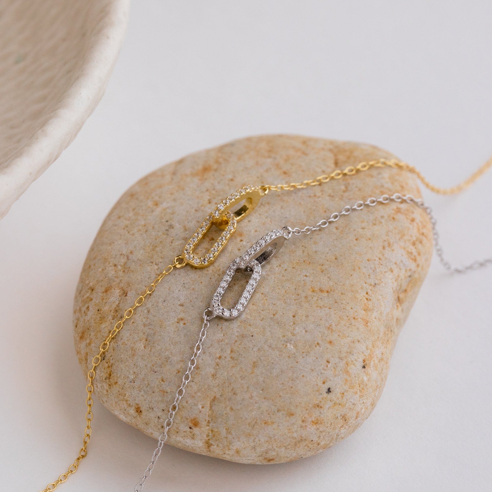 The Pave Linked Pendant Necklace, available in gold and silver with oval pendants, sparkles with pave details and glamorous style as both are elegantly displayed on a smooth stone.