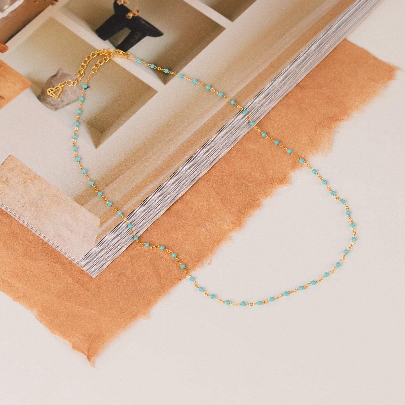 The Turquoise Station Necklace features a delicate gold chain with small turquoise beads, displayed on an open book and brown paper—perfect for a subtle boho look.