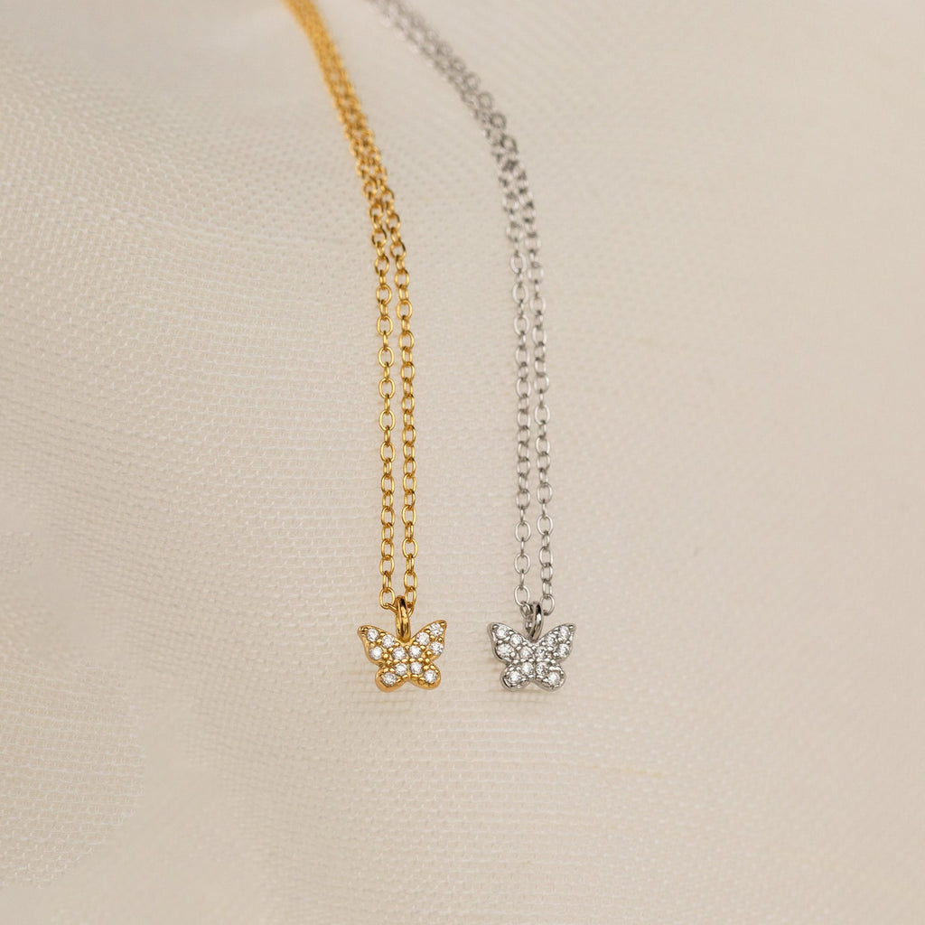 The Mandy Pave Butterfly Necklace features gold or silver pendants adorned with sparkling crystals and delicate diamond accents, beautifully displayed on a light textured fabric background.