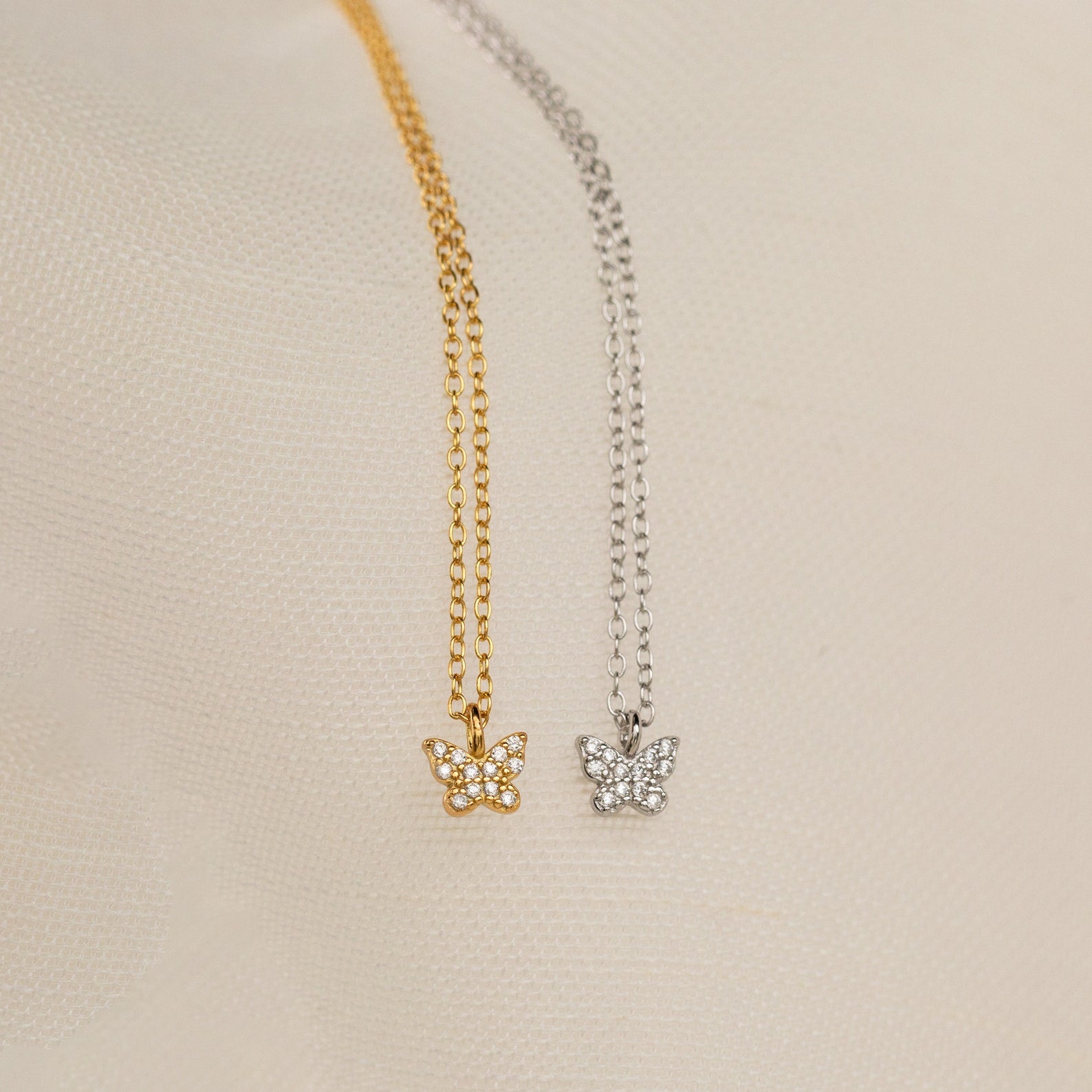 The Mandy Pave Butterfly Necklace features gold or silver pendants adorned with sparkling crystals and delicate diamond accents, beautifully displayed on a light textured fabric background.