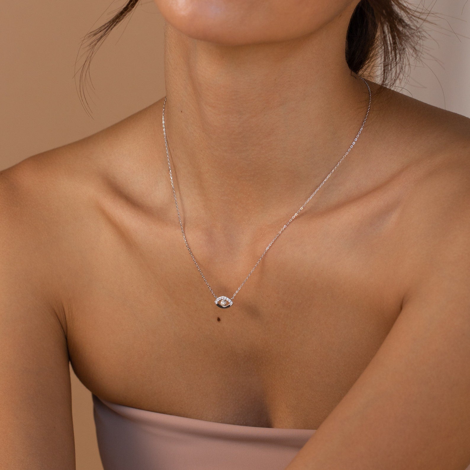 Silver necklace with an eye shaped pendant featuring a central pearl and small clear stones along the upper curve, worn close to the collarbone.