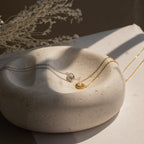 Gold and silver necklaces, including the Amaryllis Pave Evil Eye Necklace in 18K Gold, are displayed on a round textured stone dish with dried flowers in the background.