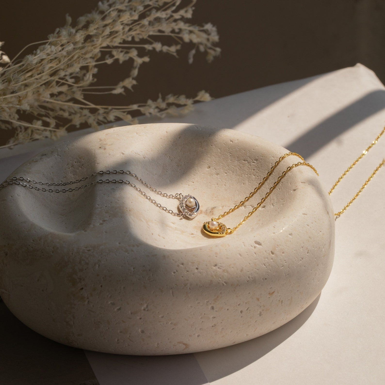 Gold and silver necklaces, including the Amaryllis Pave Evil Eye Necklace in 18K Gold, are displayed on a round textured stone dish with dried flowers in the background.