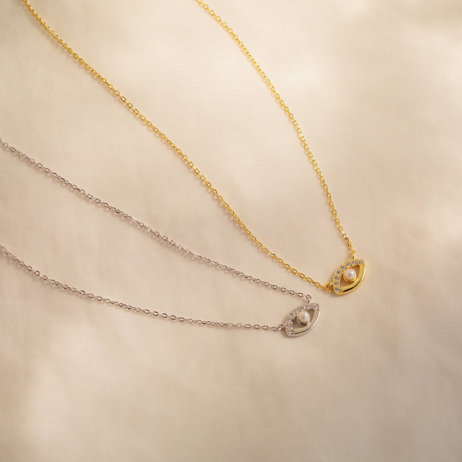 Two delicate Amaryllis Pave Evil Eye Necklaces, one gold and one silver, each featuring an oval pendant with a small pearl bead for added protection.