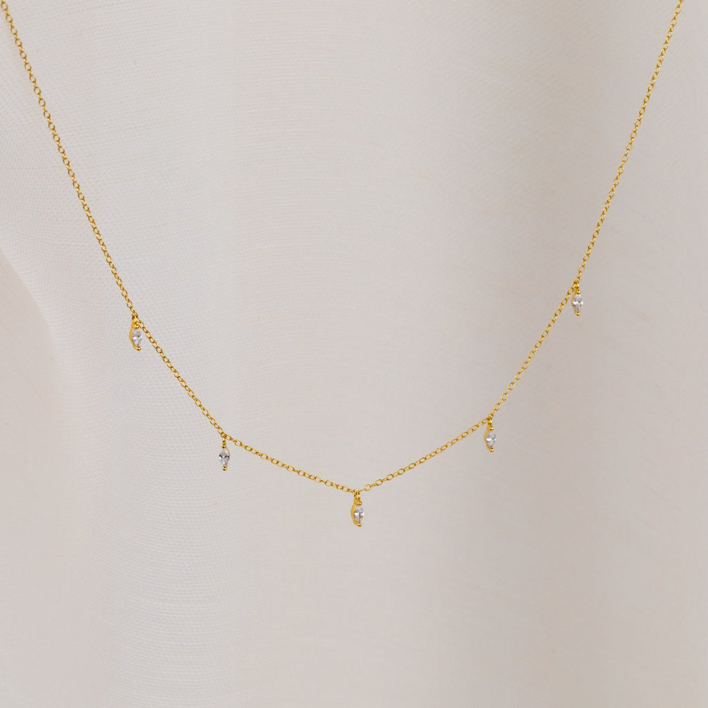 The Marquise Station Necklace features a delicate chain with small marquise-shaped diamond charms, displayed against a light fabric background.