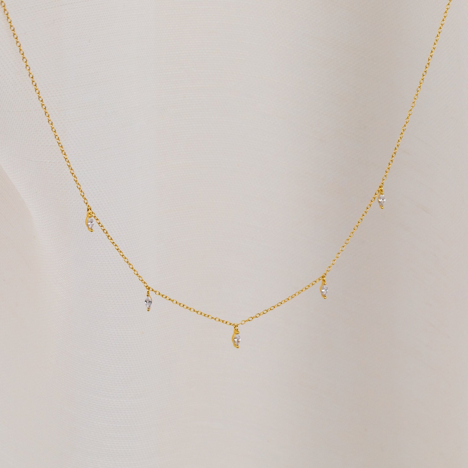 The Marquise Station Necklace features a delicate chain with small marquise-shaped diamond charms, displayed against a light fabric background.