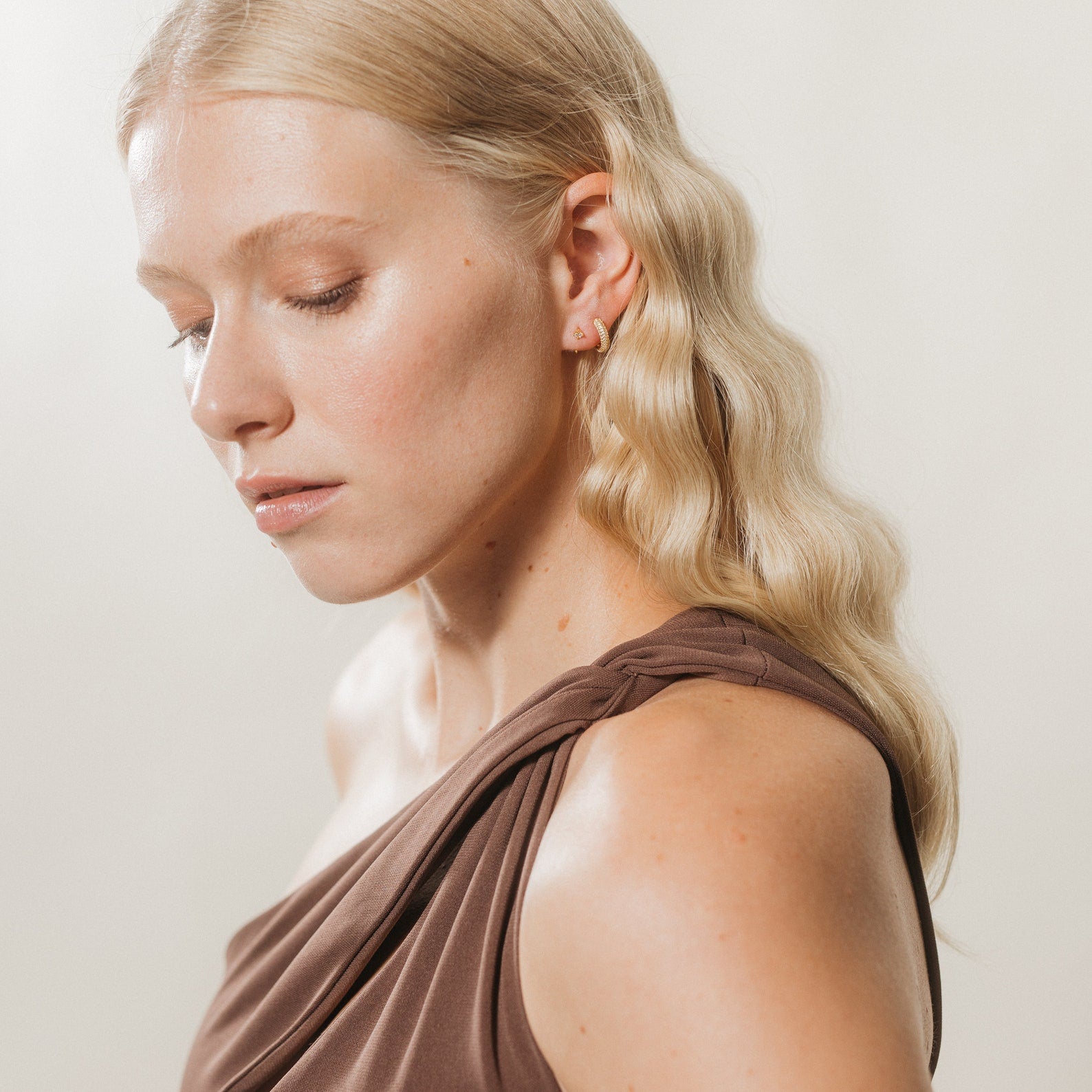 A woman with wavy blonde hair in a brown, one-shoulder top gazes down calmly, elegantly styled with Clarissa Pave Huggies—ideal date night jewelry.