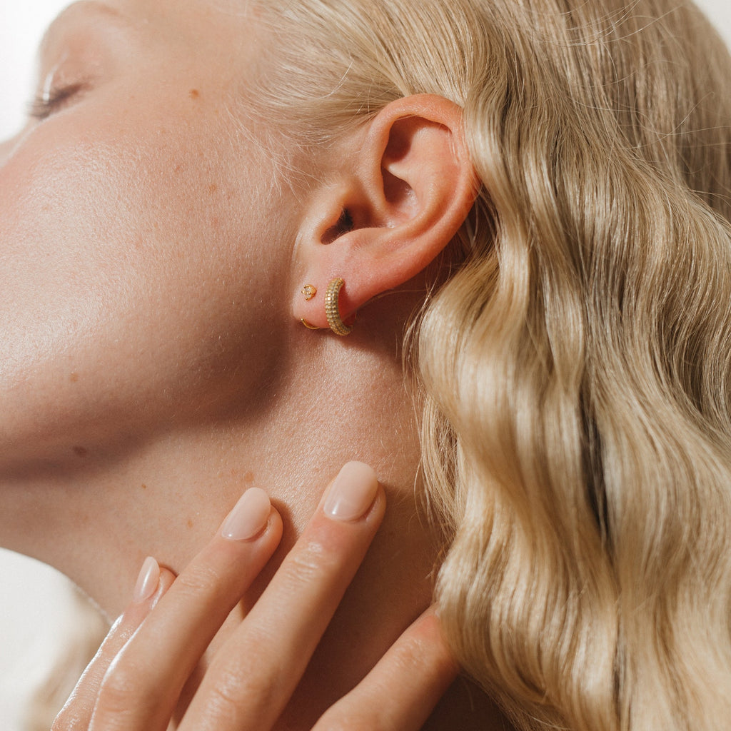 A close-up of a woman’s ear adorned with Clarissa Pave Huggies, her hand touching her neck and wavy blonde hair—a perfect date night jewelry look.
