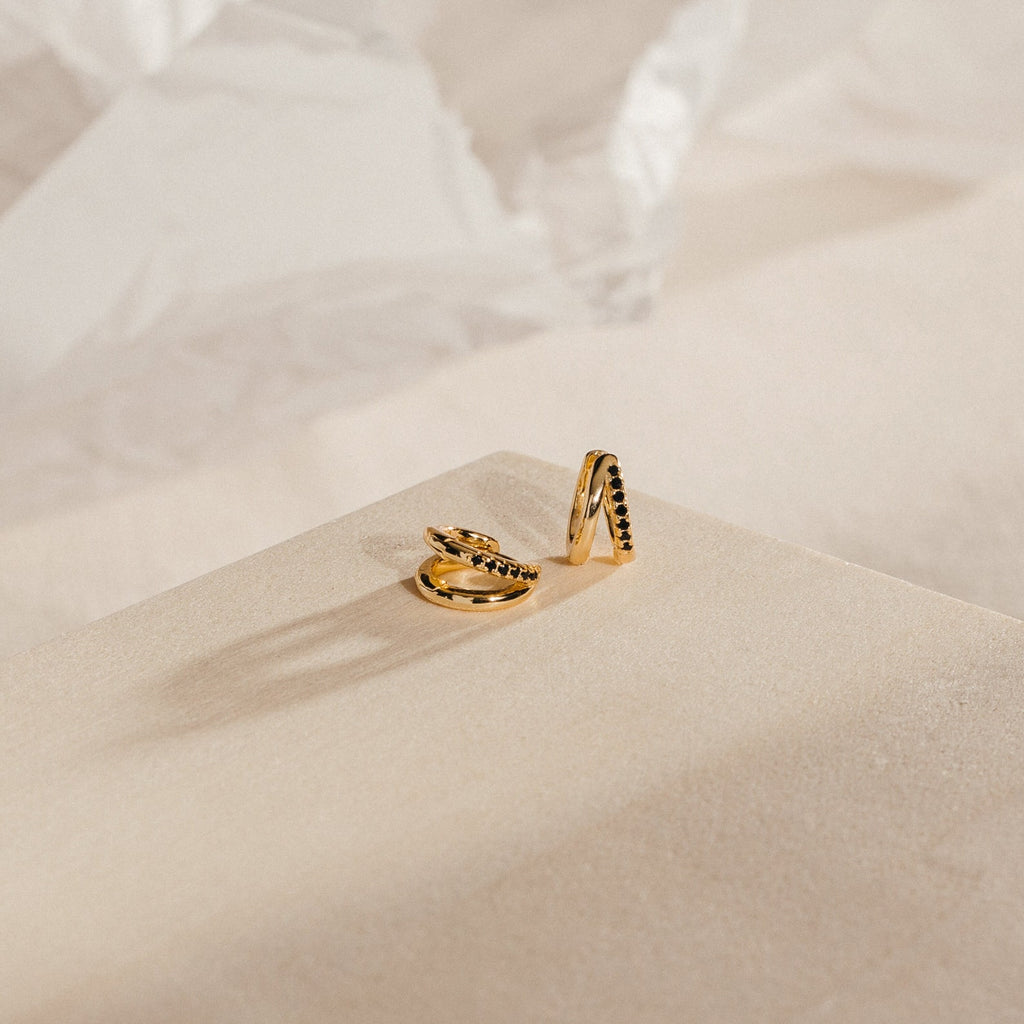 The Amelia Onyx Duo Huggies showcase gold and black stone accents on a beige textured surface, bringing minimalist elegance to any look.