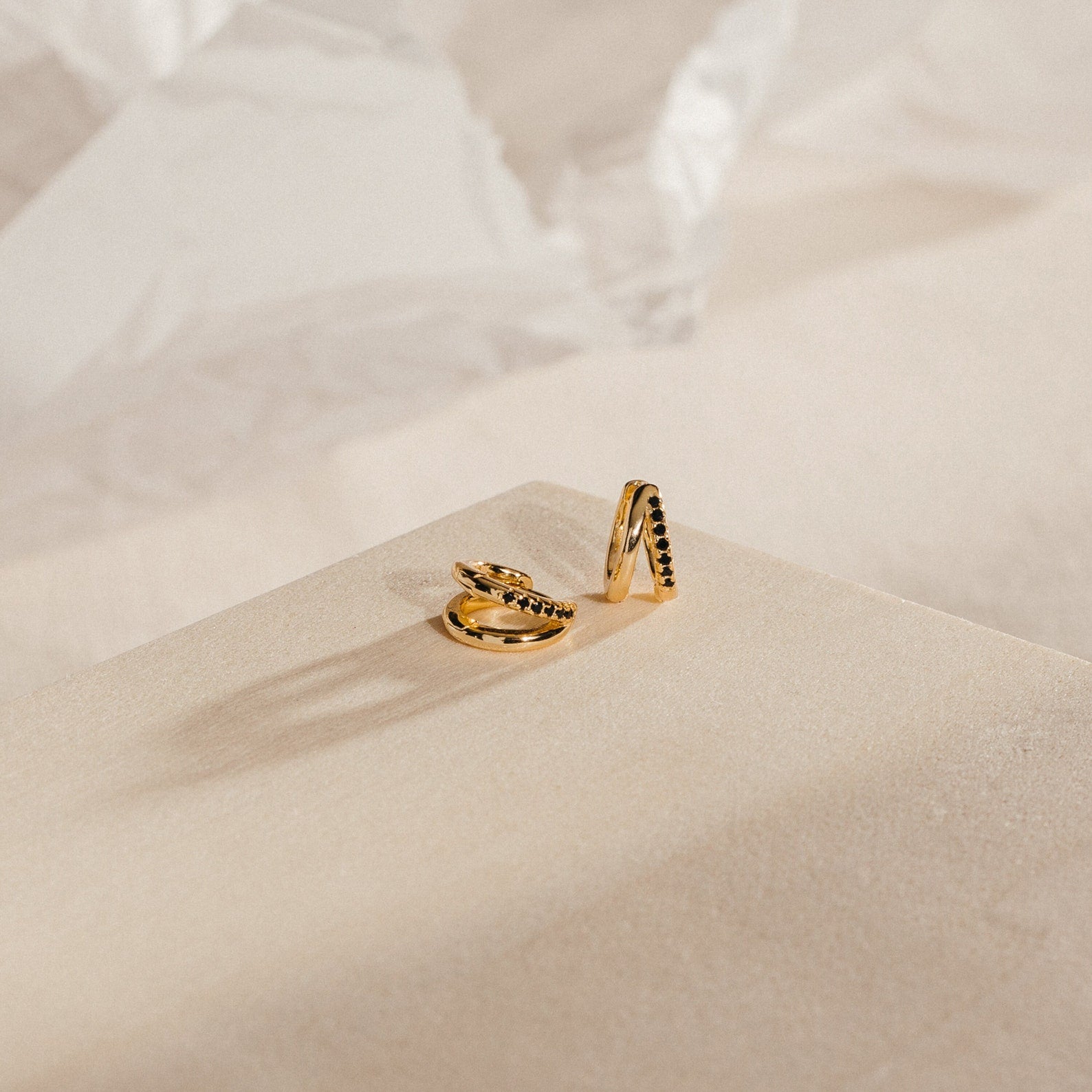 The Amelia Onyx Duo Huggies showcase gold and black stone accents on a beige textured surface, bringing minimalist elegance to any look.