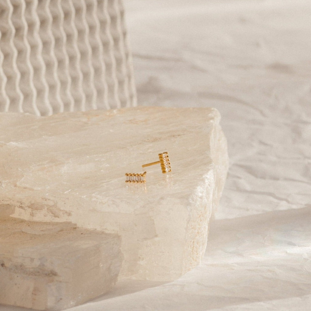 Serena Baguette Studs, elegant minimal stud earrings displayed on a textured white stone surface against a soft, neutral background.