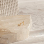 Serena Baguette Studs, elegant minimal stud earrings displayed on a textured white stone surface against a soft, neutral background.