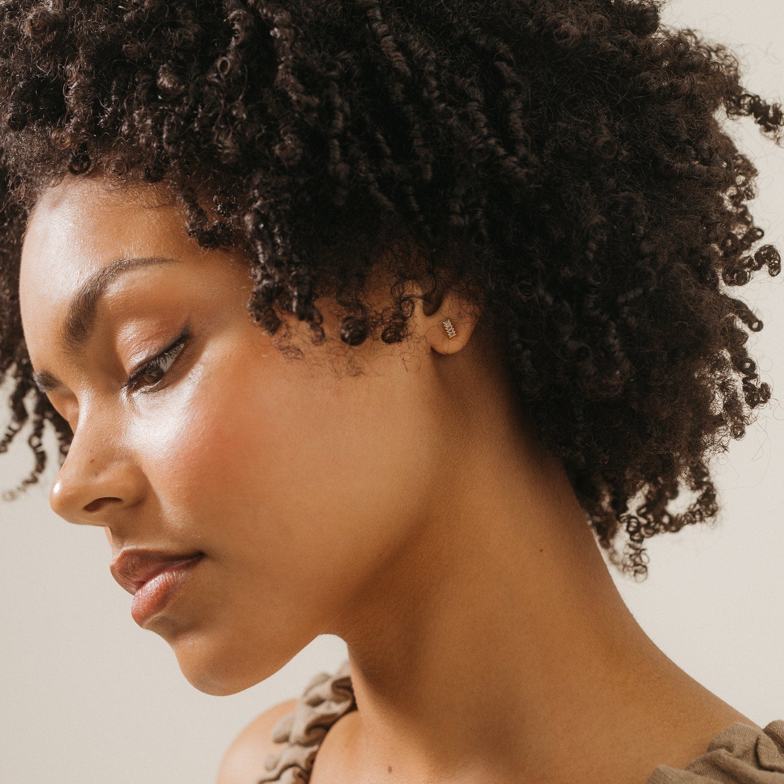 A woman with curly hair and glowing skin looks down, highlighting the Serena Baguette Studs against a neutral background.