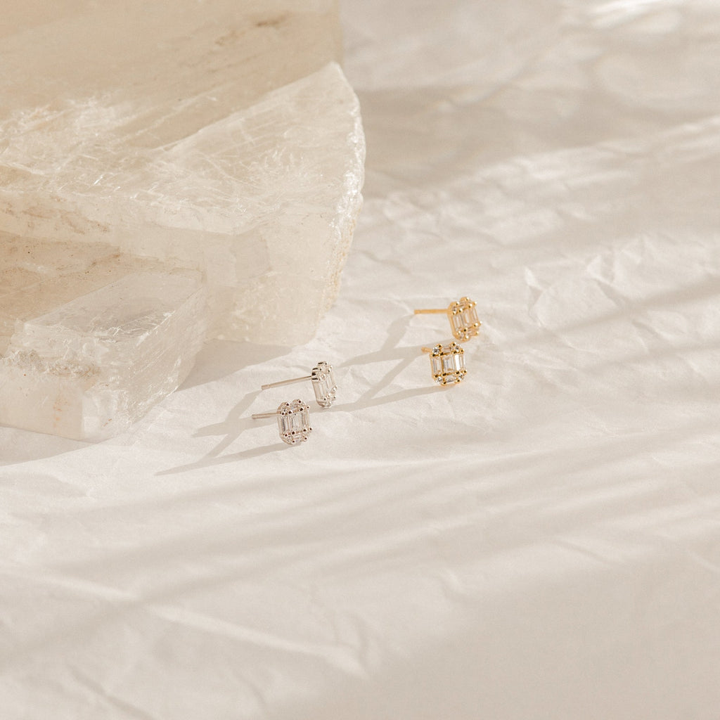 A pair of Arabella Diamond Studs rests on crumpled white paper with stone blocks, showcasing a blend of modern style and Art Deco elegance.