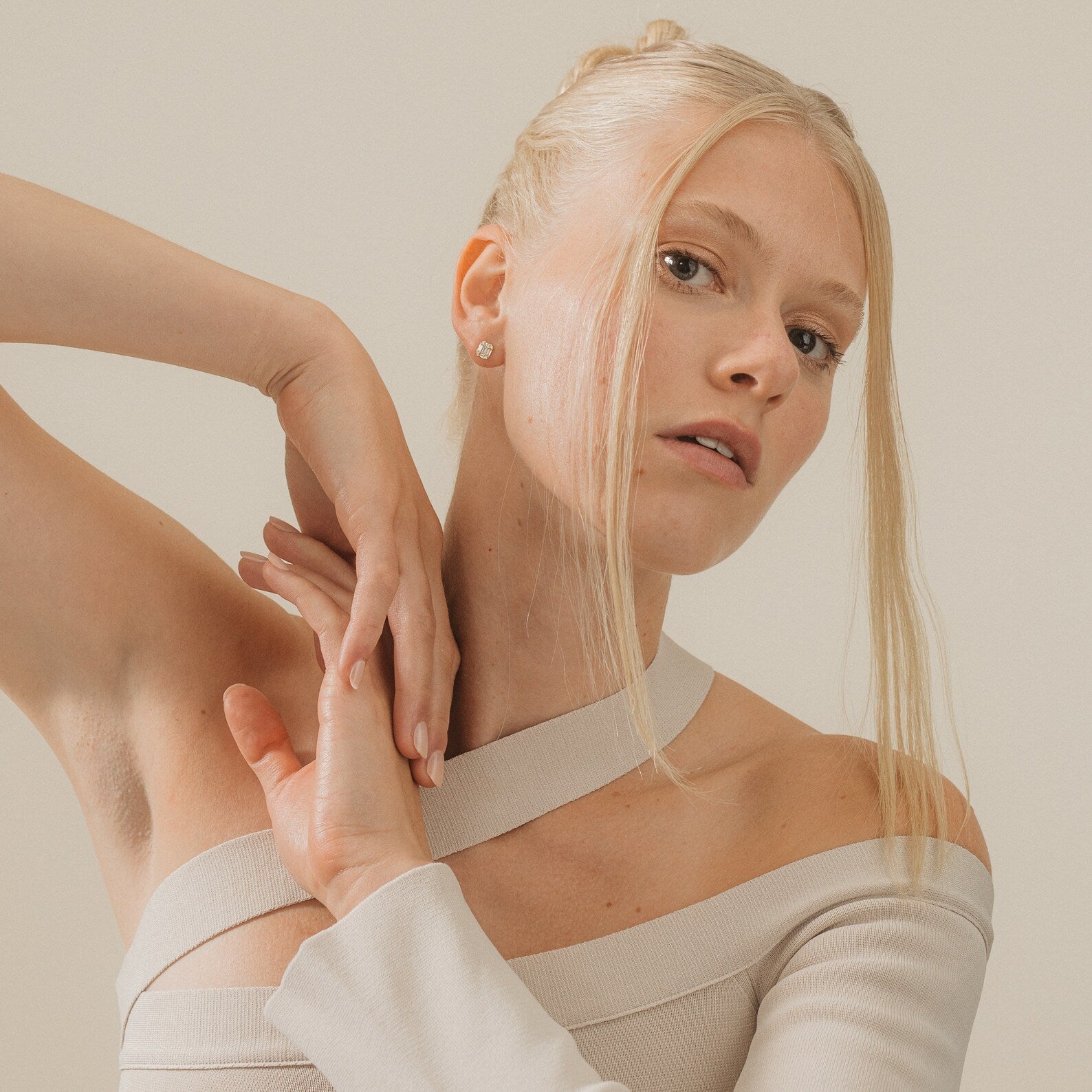 A blonde woman in a cream off-shoulder top poses with arms raised against a neutral background, showcasing the elegant Arabella Diamond Studs.