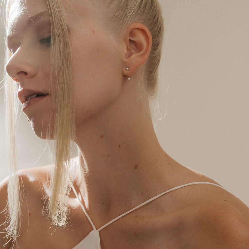 A blonde woman with fair skin in a white top gazes downward in soft natural light, elegantly showcasing Opal Ear Jackets that add a delicate accent to her serene, thoughtful style.