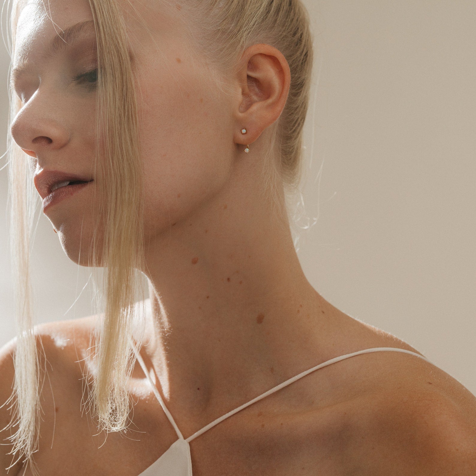 A blonde woman with fair skin in a white top gazes downward in soft natural light, elegantly showcasing Opal Ear Jackets that add a delicate accent to her serene, thoughtful style.