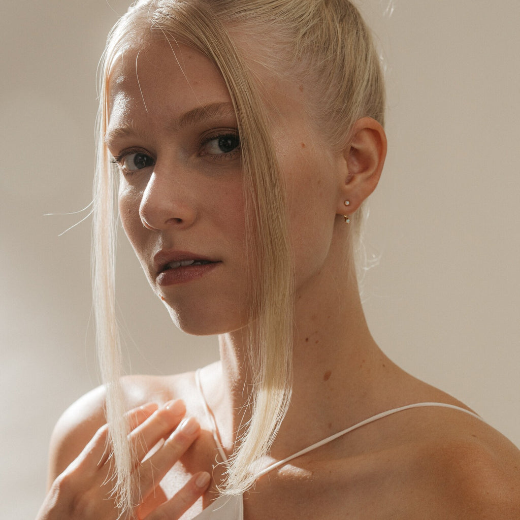 Young woman with blonde hair in a bun, two face-framing strands, wearing a white top and Opal Ear Jackets, softly lit.