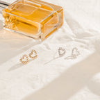 A pair of Pearl Heart Studs and a perfume bottle rest elegantly on white fabric in soft light.