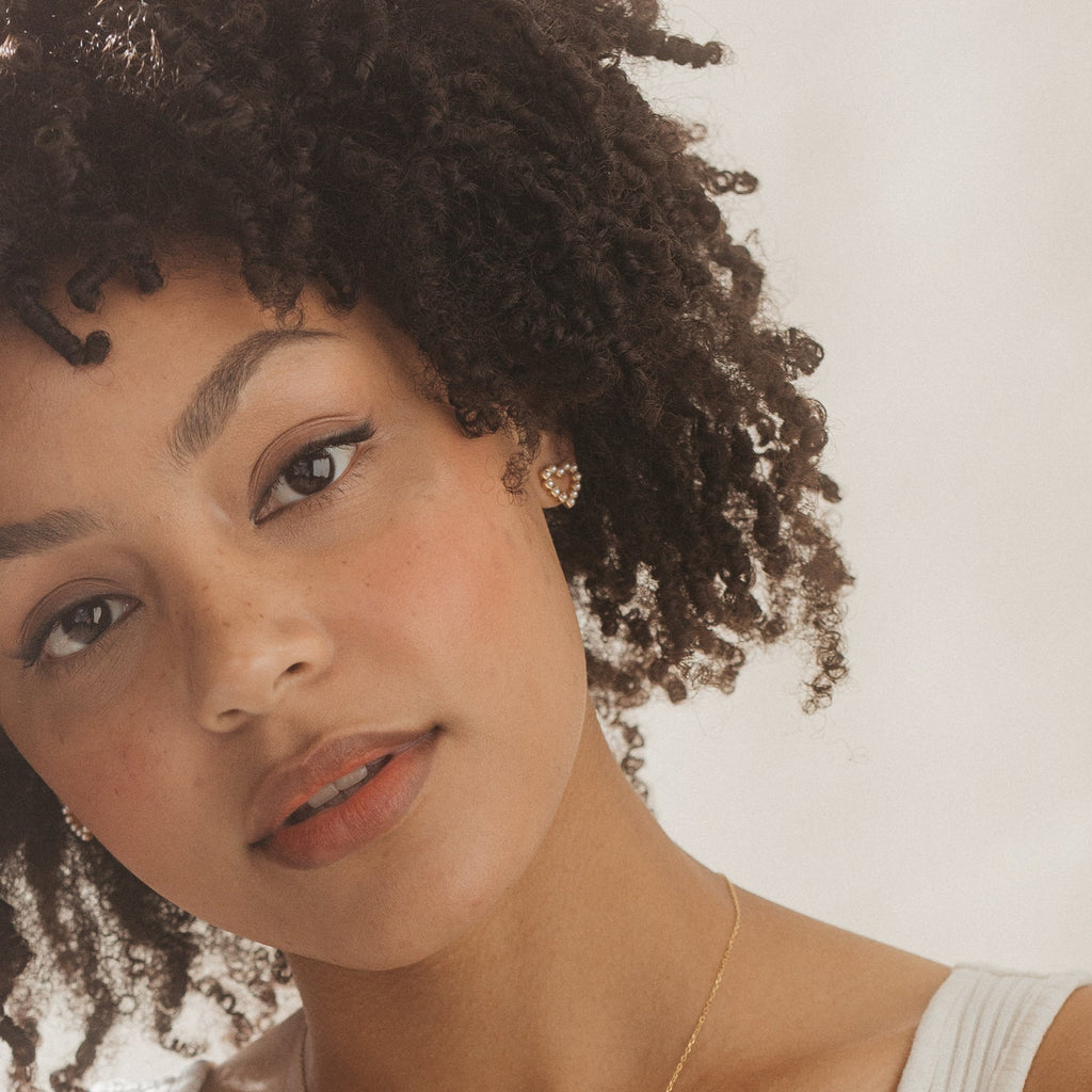 A woman with curly hair wears Pearl Heart Studs and a white top, gazing softly at the camera.