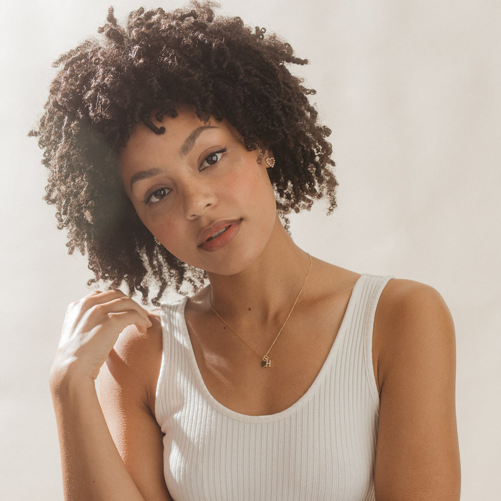 Young woman with curly hair wearing a white tank top, gold necklace, and the Pearl Heart Studs earrings, looking at the camera.