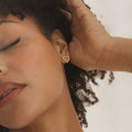 A woman with curly hair touches her ear, showcasing the Pearl Heart Studs—vintage-inspired earrings adorned with delicate small pearls.