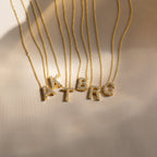 Gold necklaces featuring crystal-studded letter pendants—A, K, T, B, R, and G—are displayed on a light fabric. Elevate your look or gift someone the Floating Pave Initial Necklace for a personalized touch of sparkle.