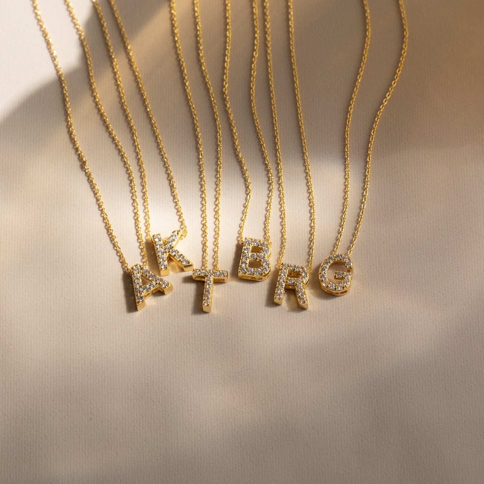 Gold necklaces featuring crystal-studded letter pendants—A, K, T, B, R, and G—are displayed on a light fabric. Elevate your look or gift someone the Floating Pave Initial Necklace for a personalized touch of sparkle.