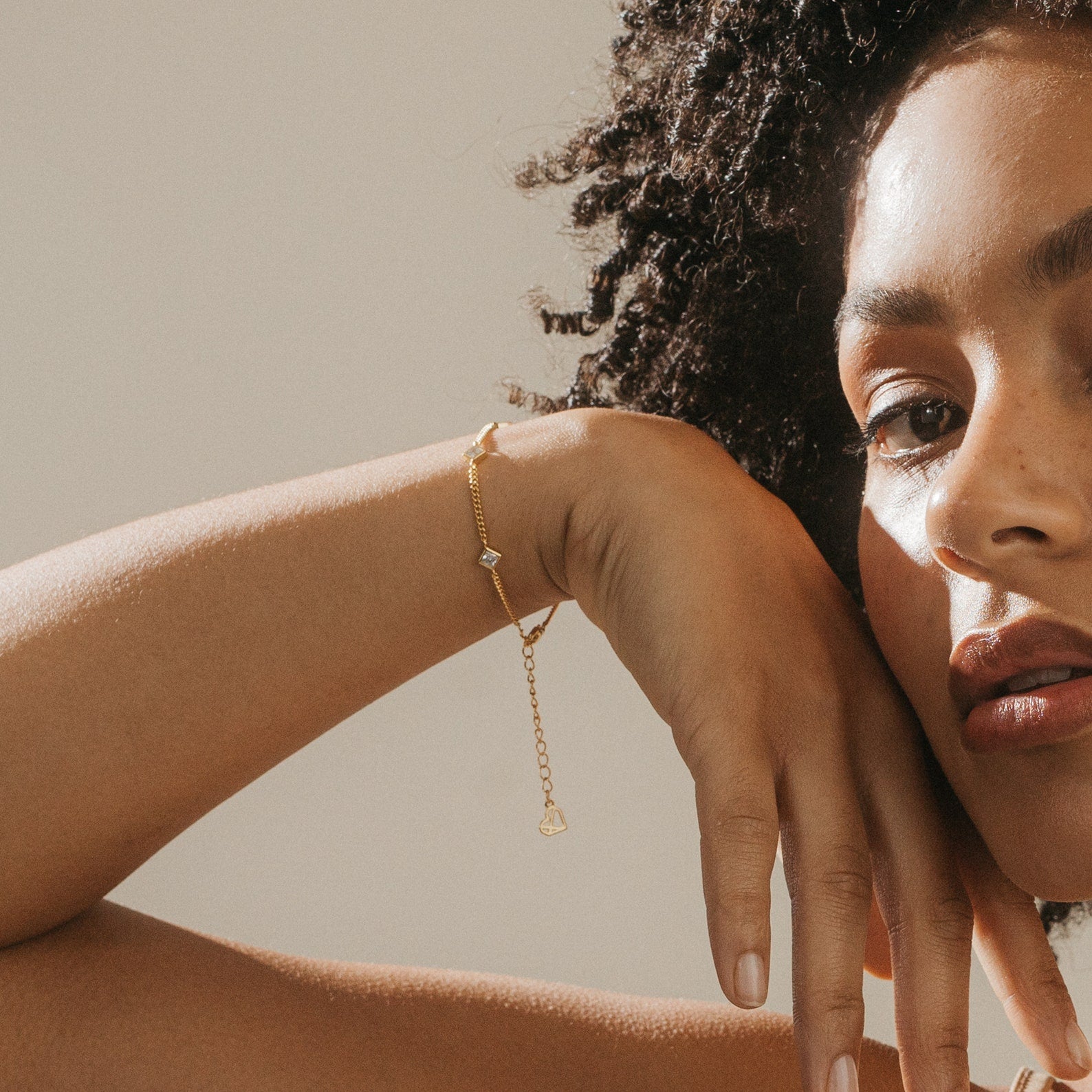 A woman with curly hair rests her hand on her face, wearing the Venus Diamond Station Bracelet—a delicate piece featuring a small charm, perfect as elegant wedding jewelry or a thoughtful anniversary gift.