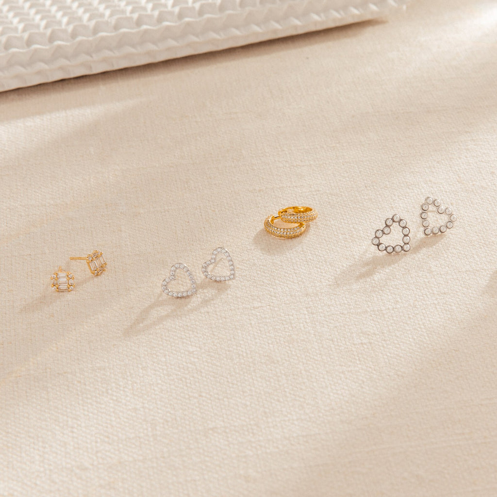 Two gold and two silver earrings, featuring bold statement styles and chic Arabella Diamond Studs, displayed on a beige textured fabric surface.