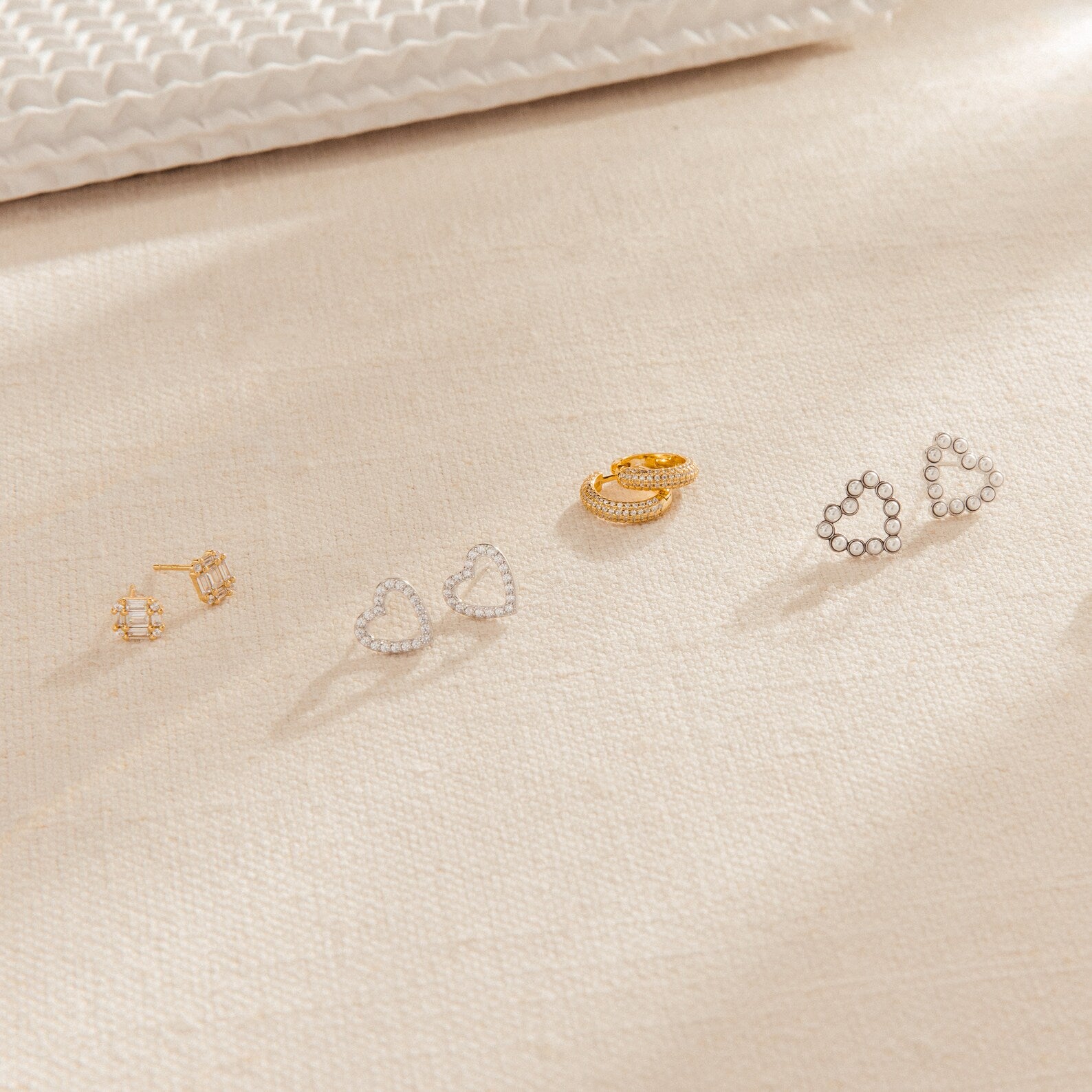 Two gold and two silver earrings, featuring bold statement styles and chic Arabella Diamond Studs, displayed on a beige textured fabric surface.