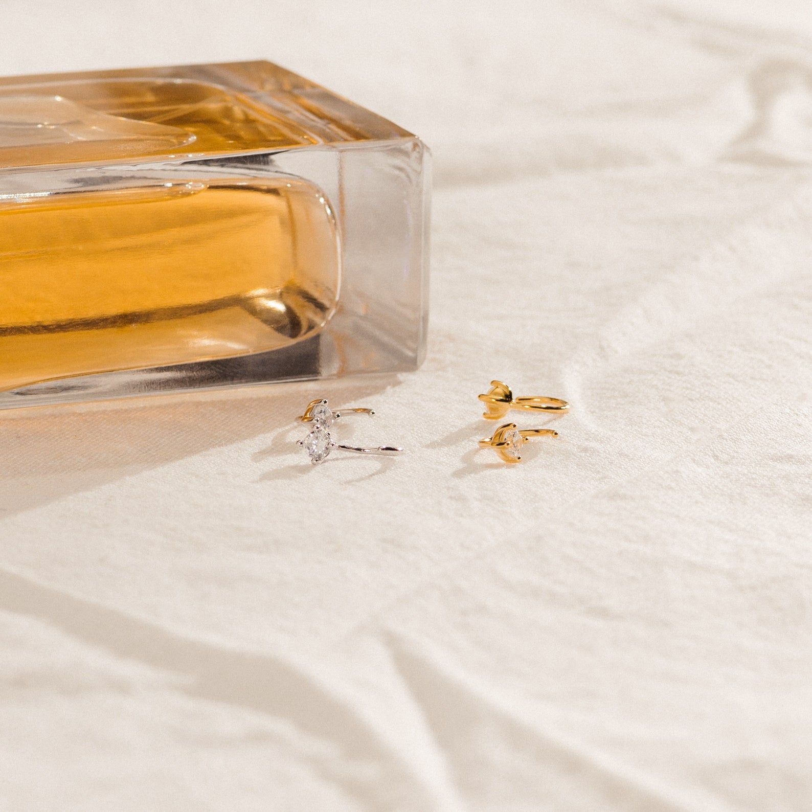A glass perfume bottle and three gold rings rest on soft beige fabric, accompanied by Saylor III Huggies—dainty hoop earrings perfect for everyday elegance.