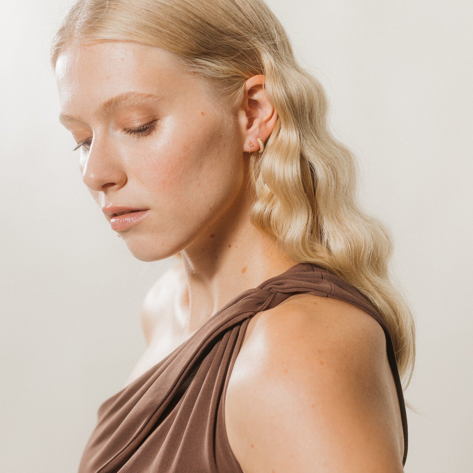 Woman with wavy blonde hair in a brown one-shoulder top looks down, highlighting Saylor II Huggies dainty hoop earrings against a light background.