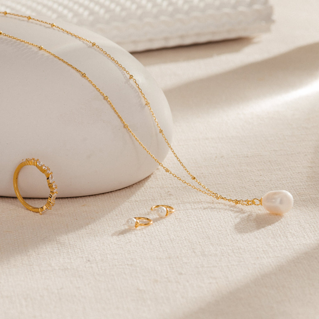 The Ariel Pearl Necklace, paired with a gold ring and pearl earrings, is elegantly displayed on a beige textured surface—ideal for bridal or bridesmaid jewelry.