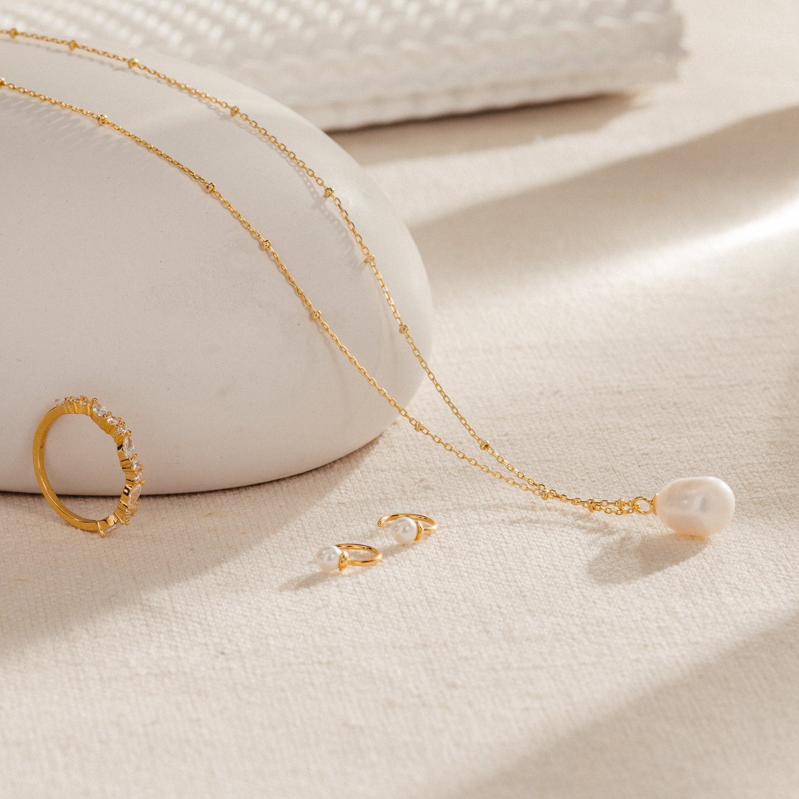 The Ariel Pearl Necklace, paired with a gold ring and pearl earrings, is elegantly displayed on a beige textured surface—ideal for bridal or bridesmaid jewelry.