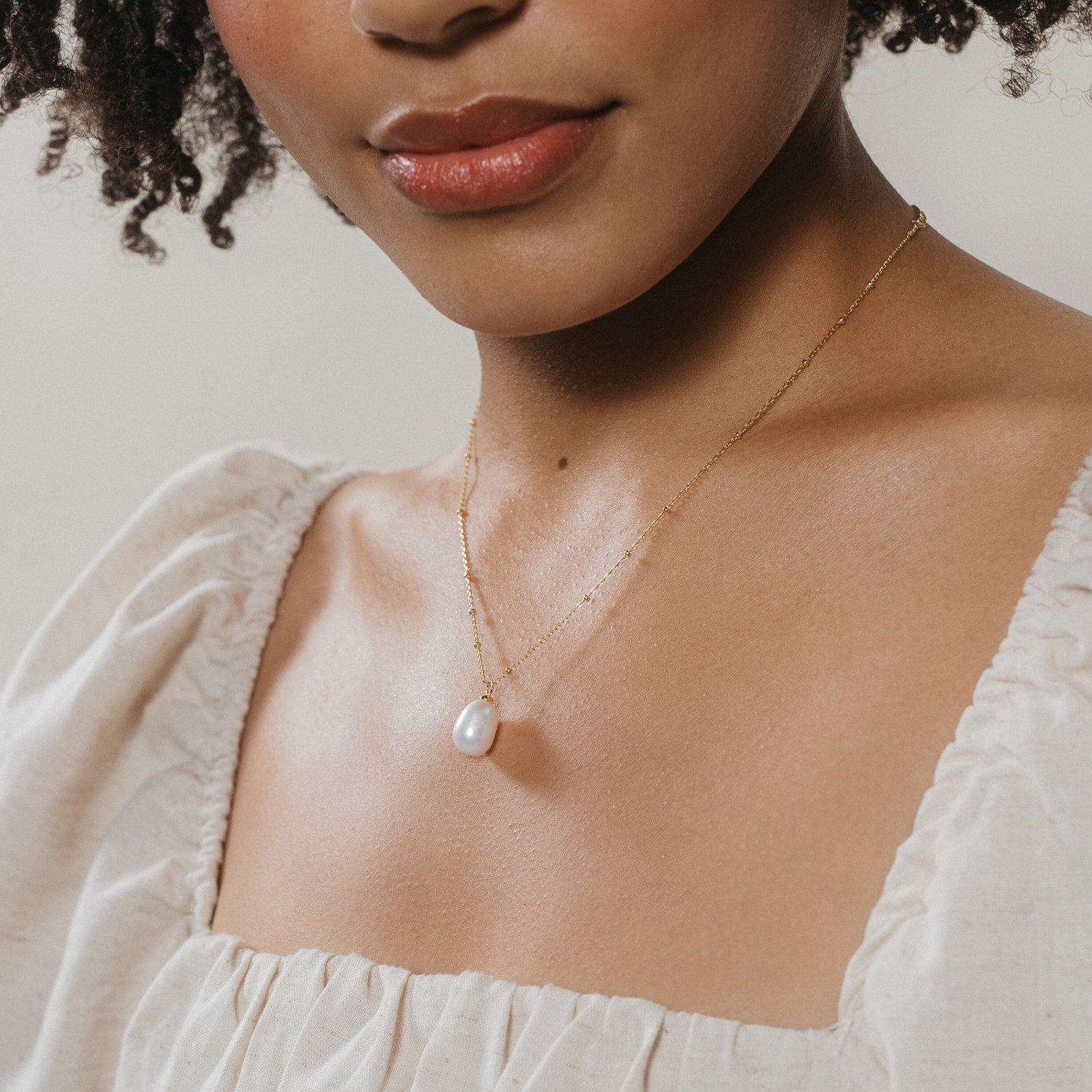Close-up of a woman wearing a gold chain necklace with a single large oval freshwater pearl pendant, styled with a cream ruched square-neck blouse.