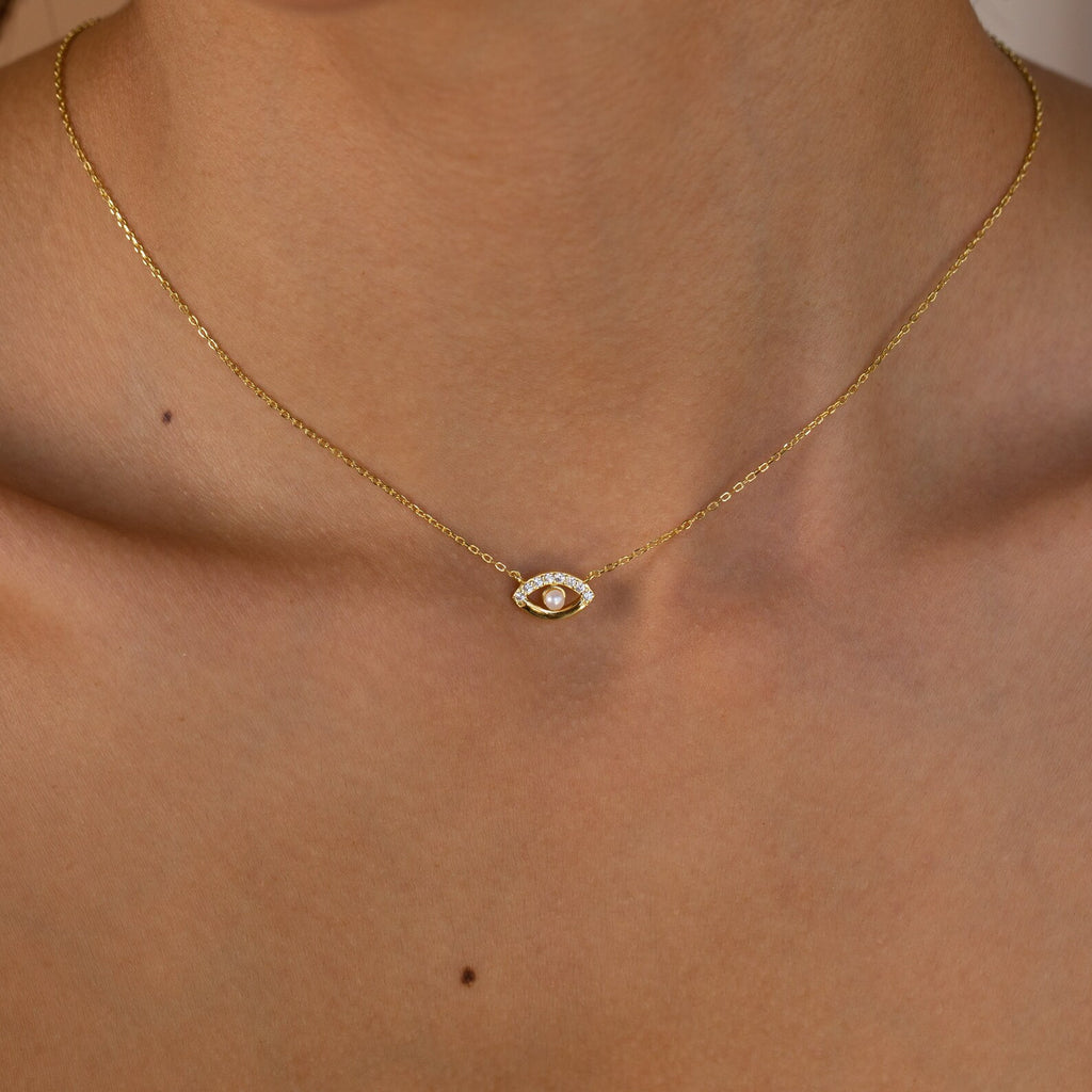 Gold necklace with an eye-shaped pendant featuring a central pearl and small clear stones along the upper curve, worn close to the collarbone.