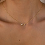 Gold necklace with an eye-shaped pendant featuring a central pearl and small clear stones along the upper curve, worn close to the collarbone.