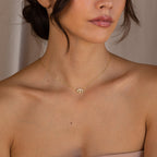 A woman wears the Amaryllis Pave Evil Eye Necklace, featuring a gold and pearl bead pendant, shown from the shoulders up against a neutral background—symbolizing style and protection.