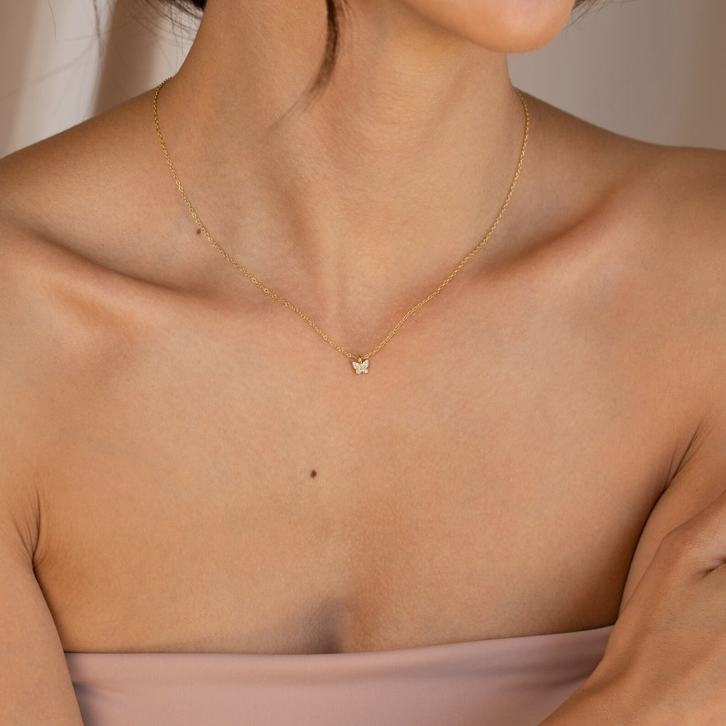 A woman wears the Mandy Pave Butterfly Necklace, featuring a delicate gold chain with a small diamond-adorned butterfly pendant, on her bare neck and shoulders.