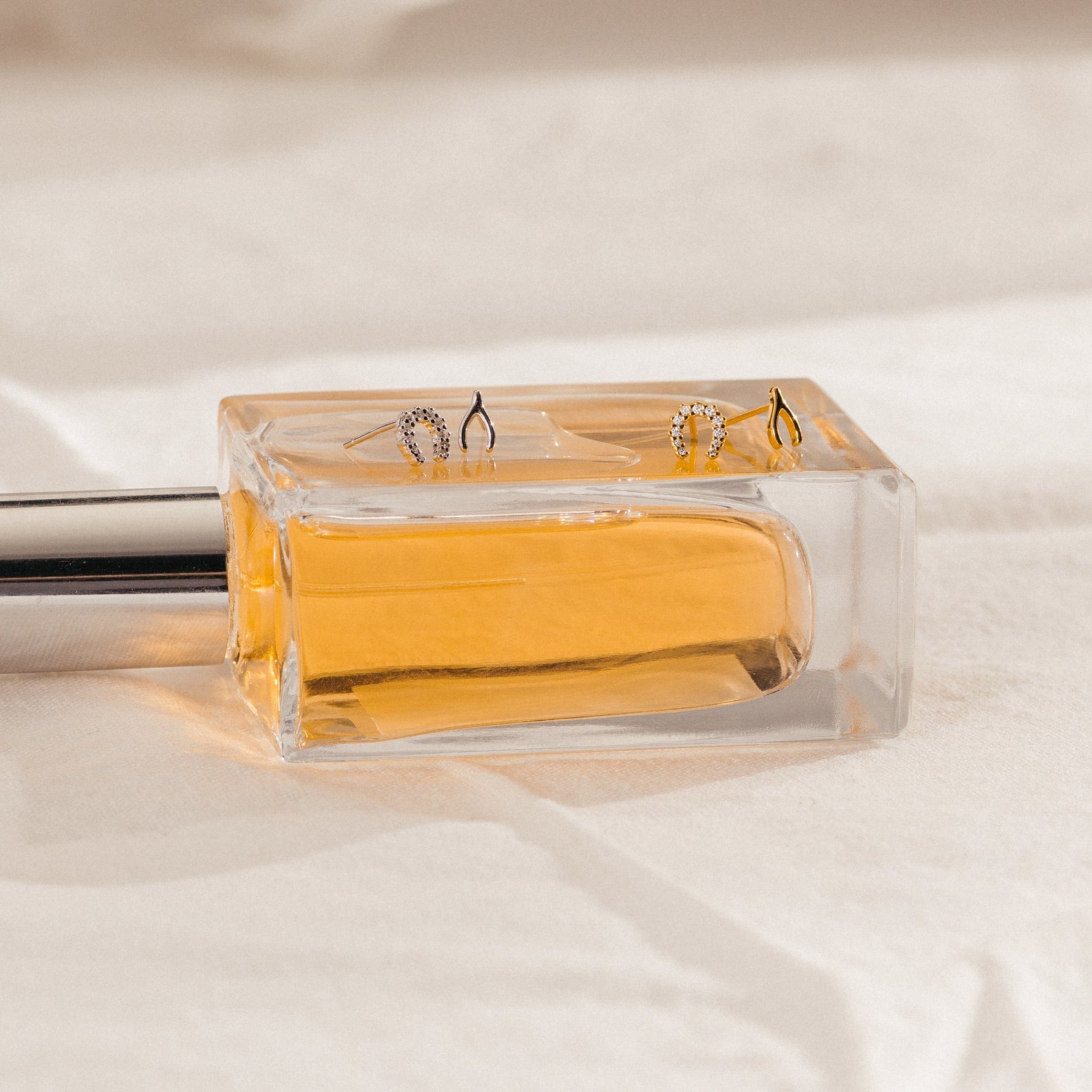 Three delicate rings rest atop a clear rectangular perfume bottle filled with amber liquid, next to the Lucky Studs Set—a collection of mismatched stud earrings that lend an artful touch to the scene.