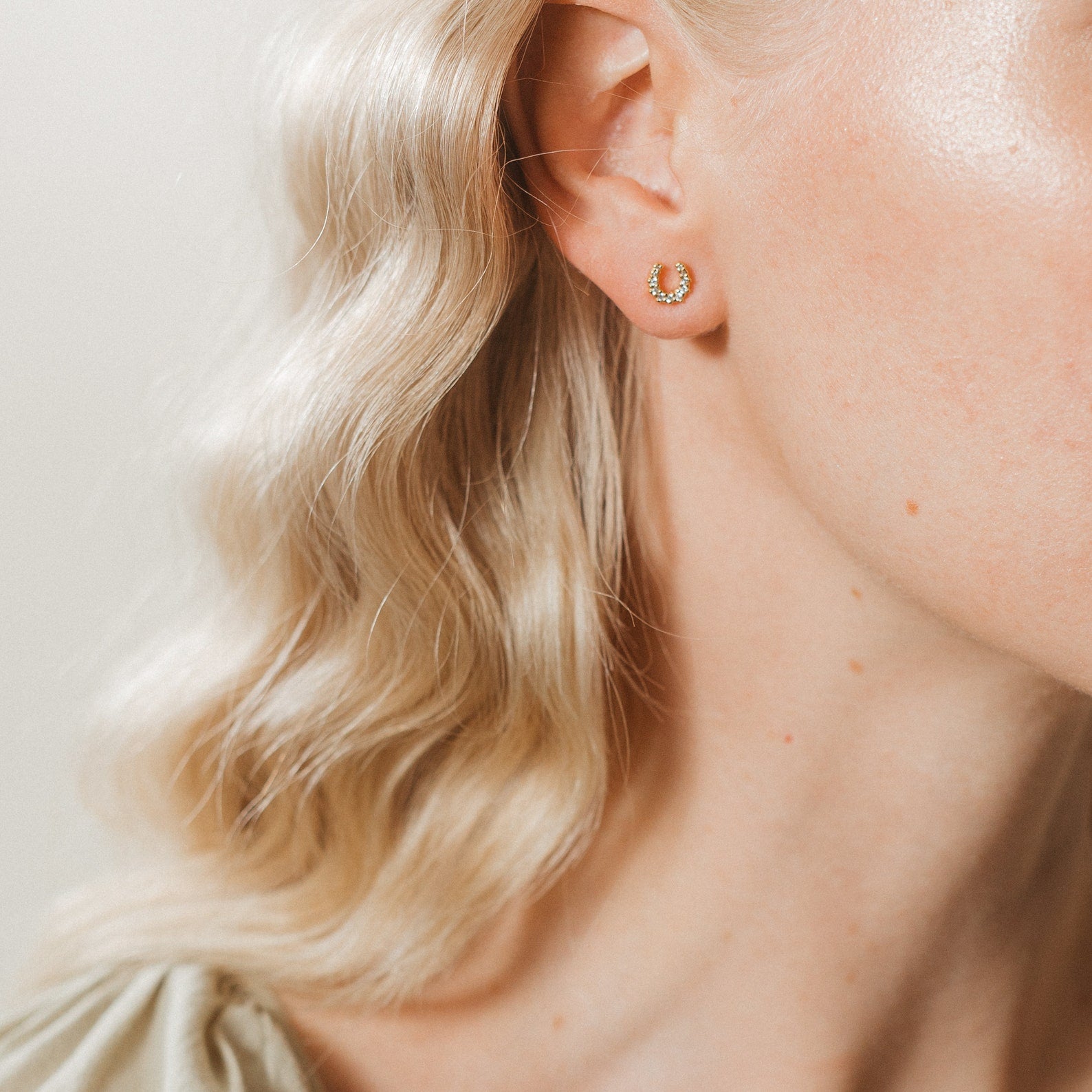 A close-up of a woman's ear with wavy blonde hair showcases the Lucky Studs Set: a small gold hoop earring with green stones paired with a delicate, mismatched stud.