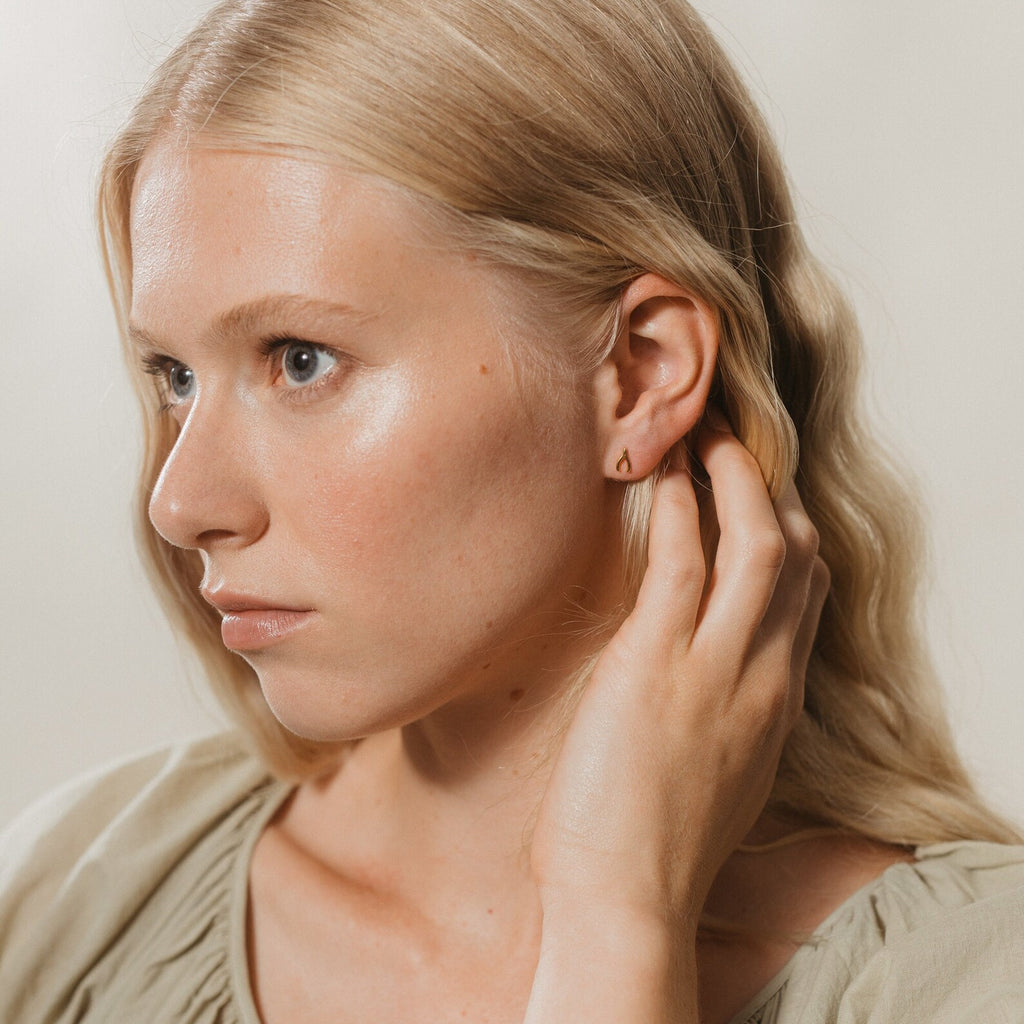 A woman with blonde hair touches her ear, wearing the Lucky Studs Set—mismatched stud earrings including a small gold piece—as she looks to the side.