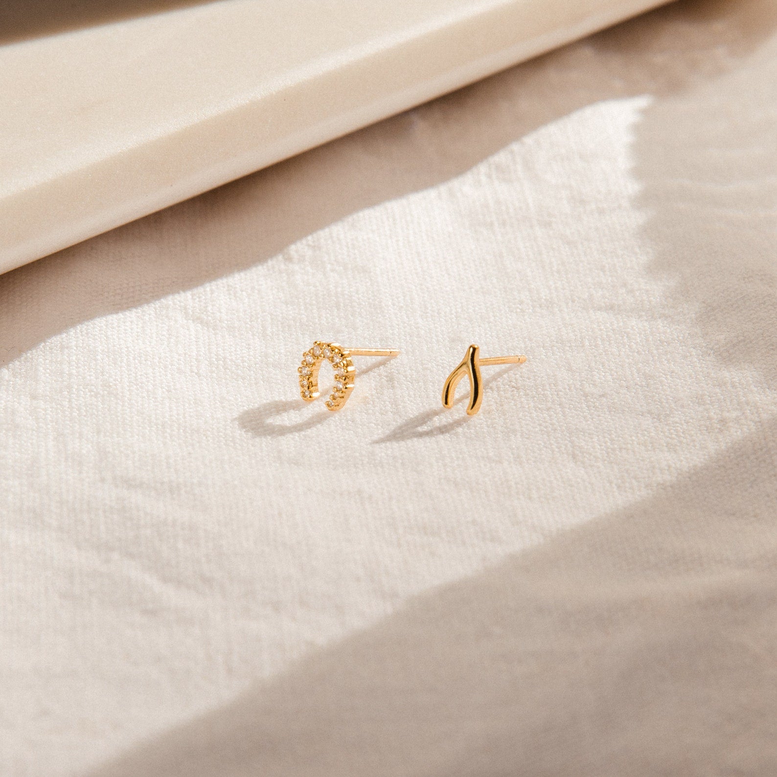 The Lucky Studs Set, featuring a wishbone and a diamond horseshoe stud earring, rests on a softly sunlit fabric surface.