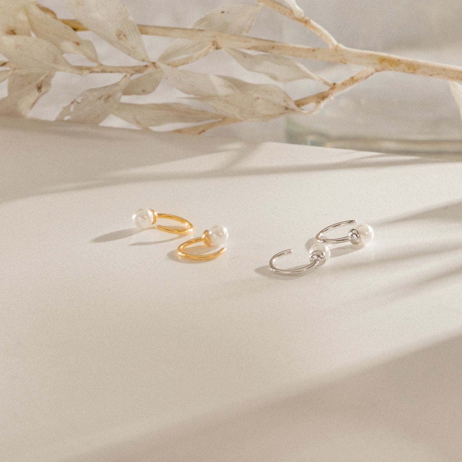 The Lila Pearl Huggies—dainty hoop earrings in gold and silver, each adorned with pearls—are displayed on a white surface with dried leaves, offering the perfect touch of modern jewelry elegance.