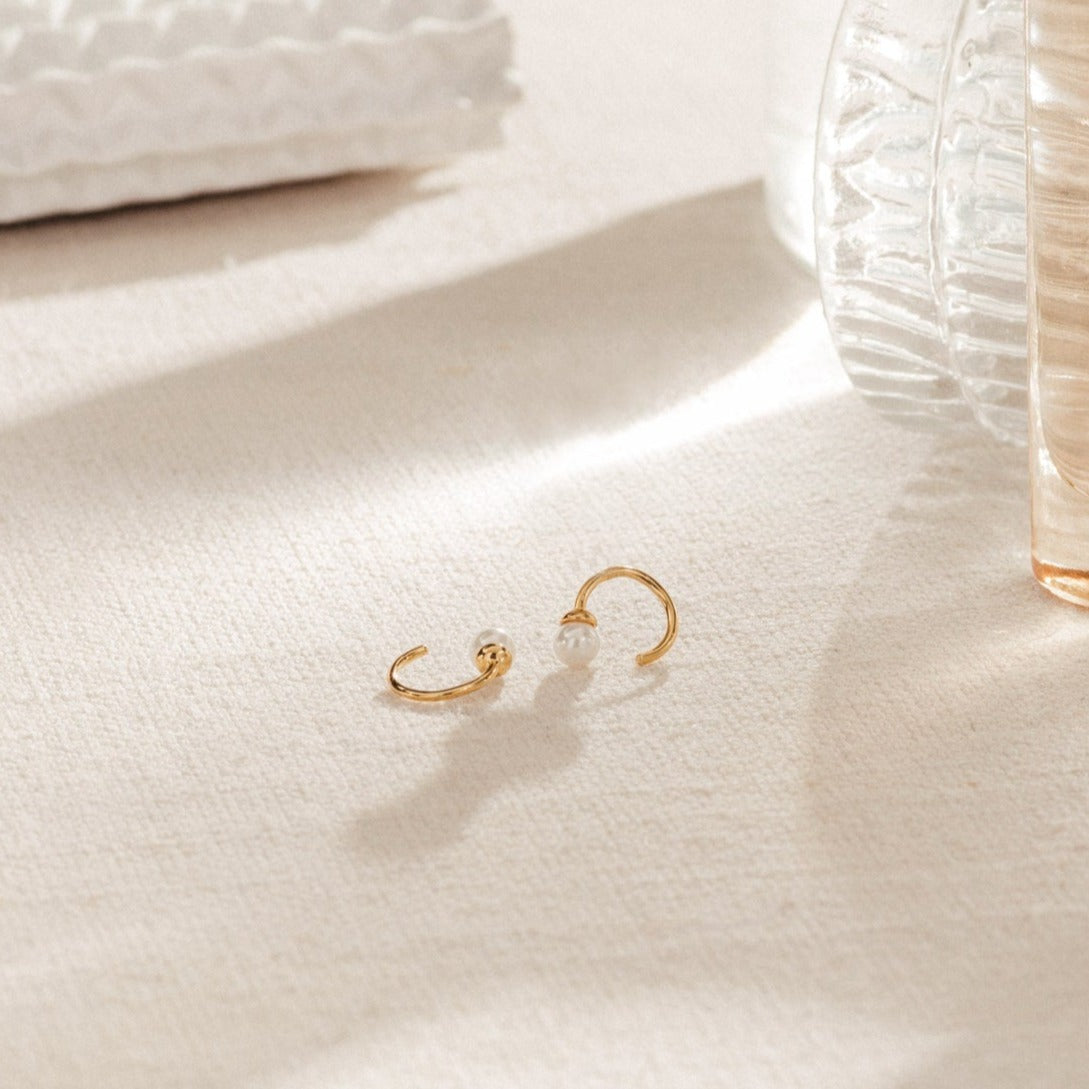 The Lila Pearl Huggies, featuring dainty gold hoops with pearl and bead accents, rest on a textured surface in soft light—ideal for lovers of modern jewelry.