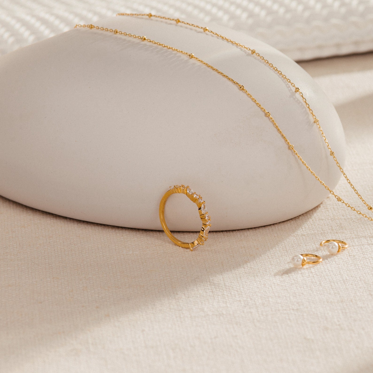 The Verity Diamond Ring, gold necklace, and pearl earrings are displayed on a smooth, beige surface.